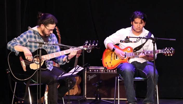 A teenage boy and his teacher seated together, both playing electric guitars on stage during a recital