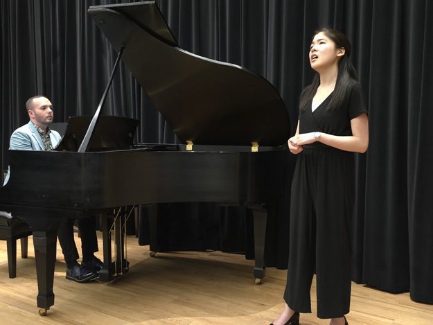 An adult female performs a classical song at a recital, standing poised on stage with her voice teacher accompanying her at the piano