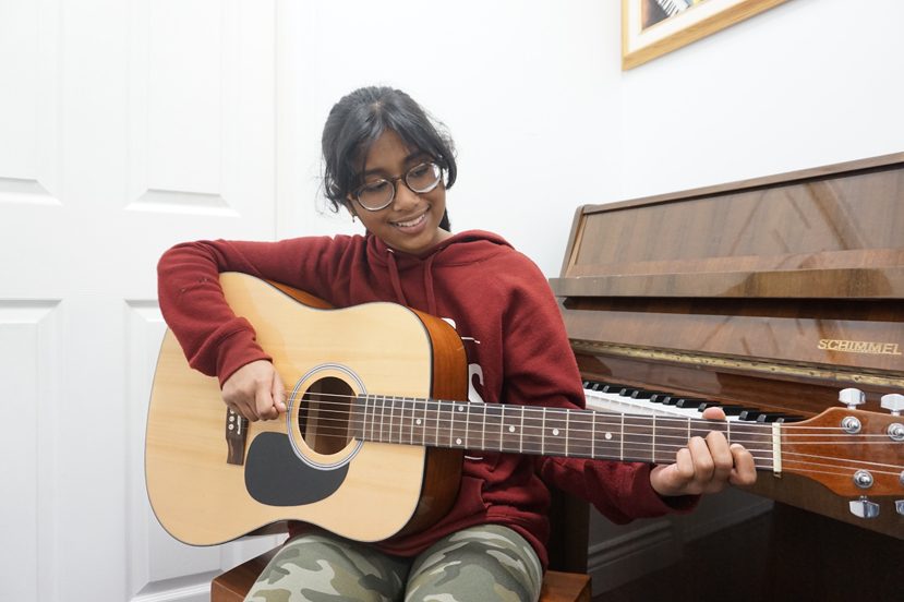 Young girl playing acoustic guitar in front of a Schimmel piano