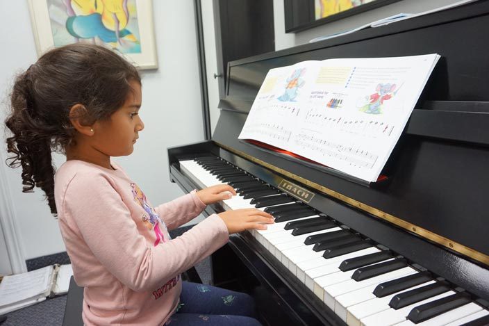 A little girl in Mississauga sitting at an Ibach upright piano during her piano lesson, playing from a children's music book