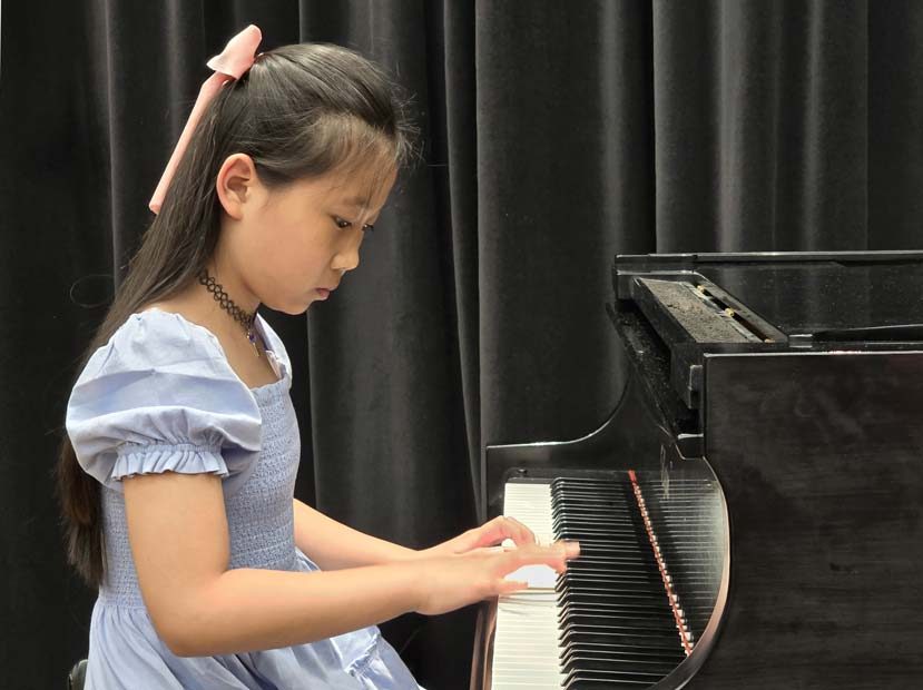 A young girl in a blue dress plays a classical piece at a grand piano recital, focused and poised
