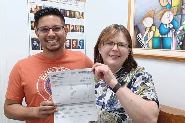 An adult male piano student and his teacher standing together, proudly holding up his piano exam results